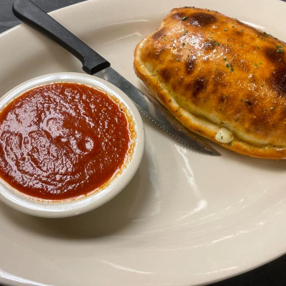 Golden-brown calzone with herbs, a side of marinara sauce, and a knife on a white plate.